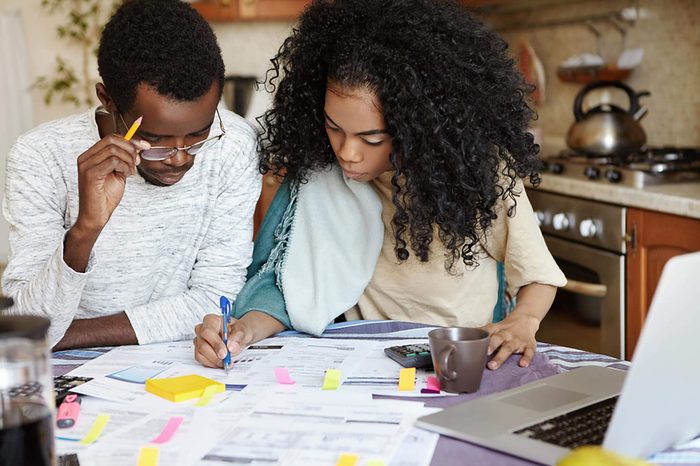 Young confident African housewife with Afro hairstyle helping her husband to manage domestic finances, calculating and making notes with pen, both sitting at kitchen table with laptop and papers