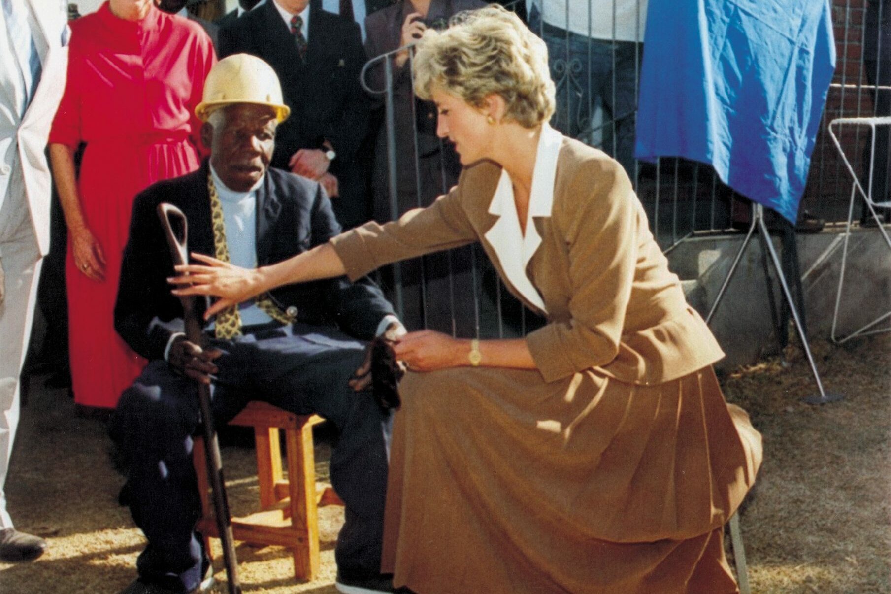A woman kneels to interact with a seated man holding a cane, surrounded by smiling onlookers in an outdoor setting.