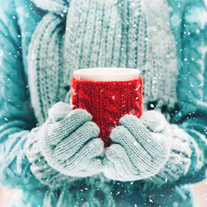 Hands hold a red mug wrapped in a knitted cover, wearing blue gloves, as snow falls gently in a cozy, wintry setting.