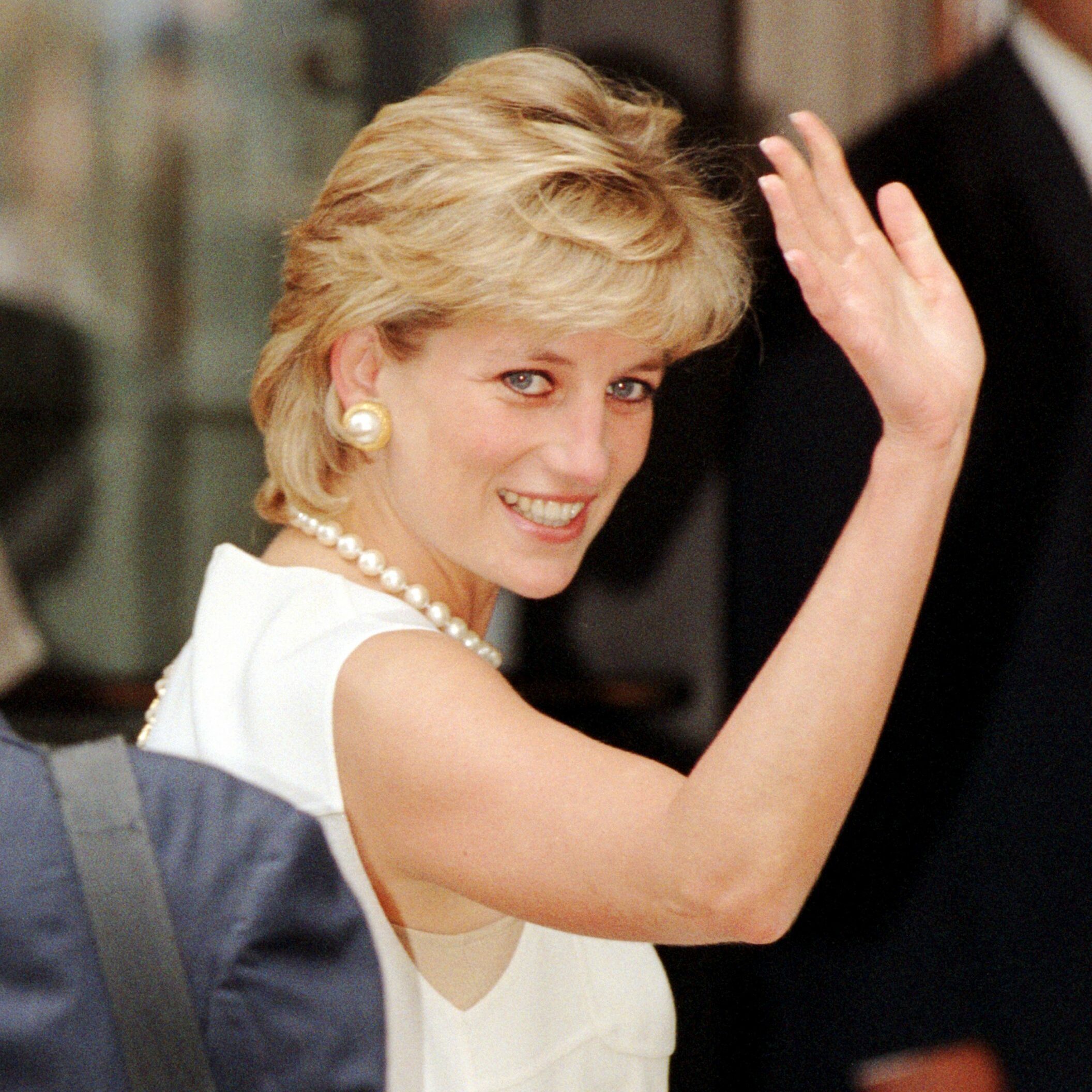 A woman in a white dress waves, smiling, in a crowd with blurred figures in the background.