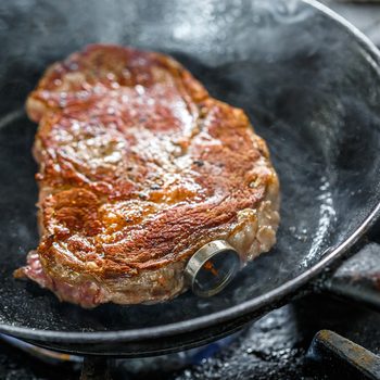 A steak sizzles in a frying pan, releasing steam, on a stove top.