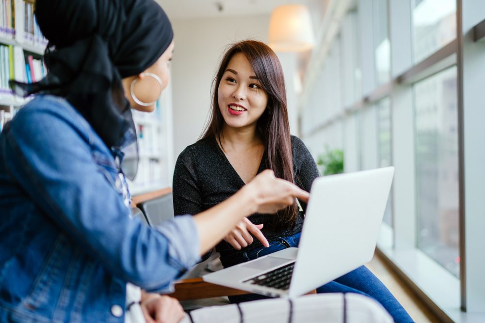 A young and attractive Chinese Asian woman has a business meeting with a Malay Muslim woman. They are both sitting in an office and talking over a laptop computer during the day. 