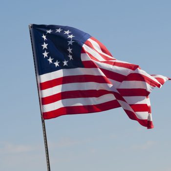 A flag with thirteen stars in a circular pattern waves against a clear blue sky.