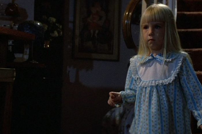 A young girl stands in a dimly lit room, wearing a blue nightgown, with a staircase visible in the background.