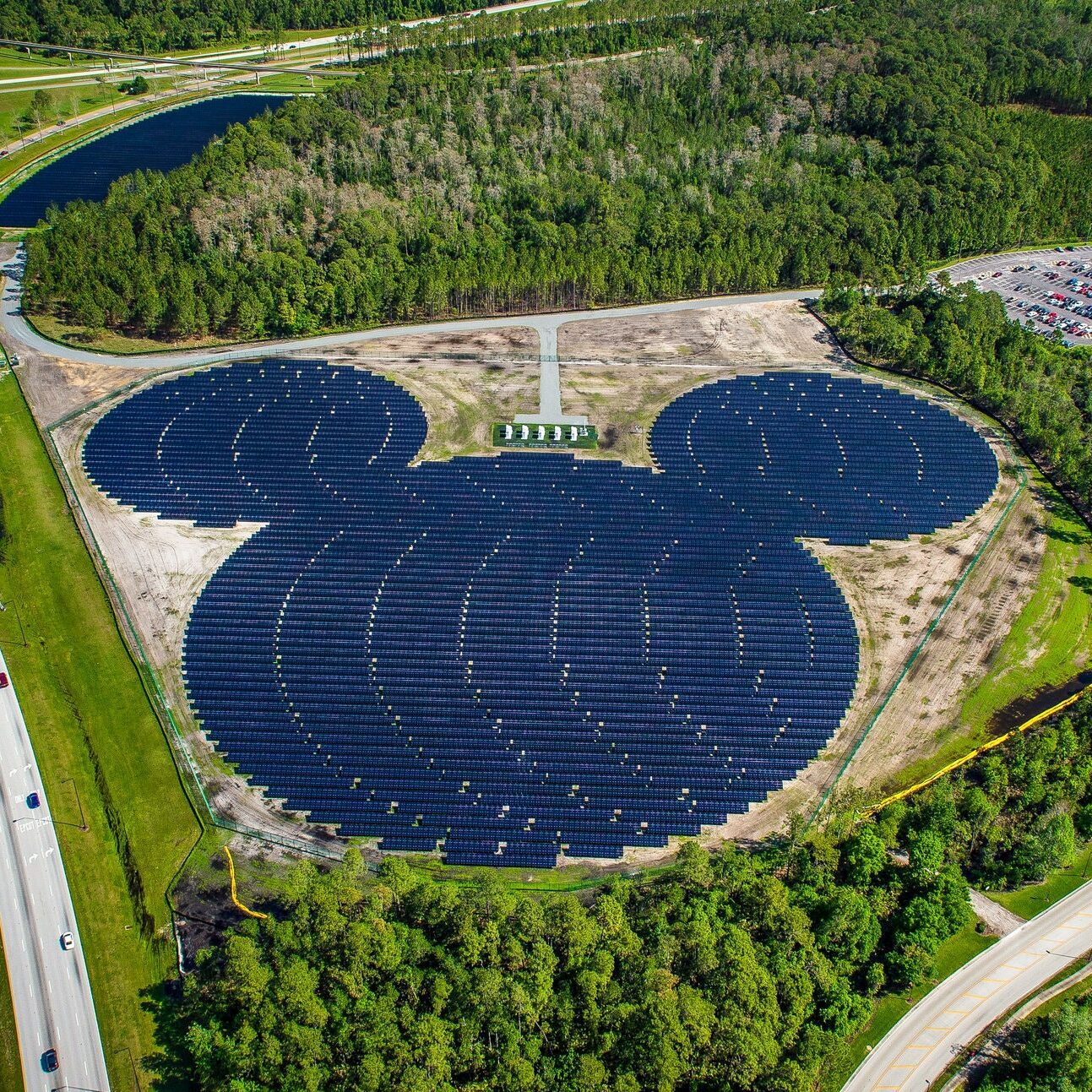 Solar panels form a Mickey Mouse shape; surrounded by greenery, parking lot, and roads in a landscaped area.