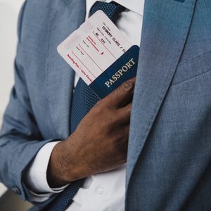 Hand holding passport and boarding pass, placing them in suit pocket; context includes a coffee cup and smartphone on a desk.
