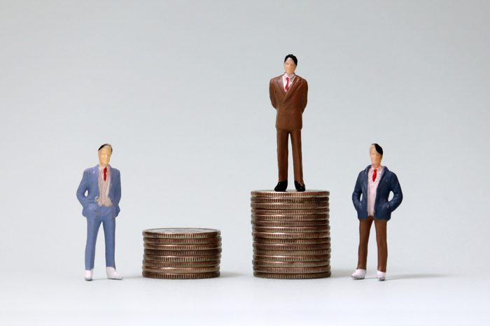 Two miniature men standing next to a miniature man standing on a pile of coins.