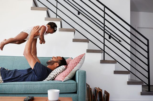 Man lifts baby joyfully on a teal couch, beneath a modern staircase, with cushions and a coffee table nearby, creating a playful family scene.