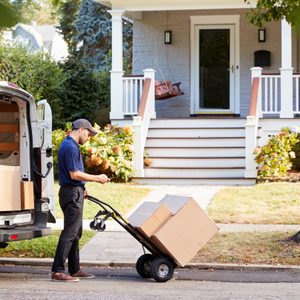 Delivery person pushes boxes on a dolly from a van towards a house with a porch and swing in a suburban neighborhood.