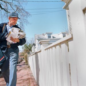 Mail carrier walks along a sidewalk, holding letters and magazines, next to a white picket fence in a residential neighborhood.