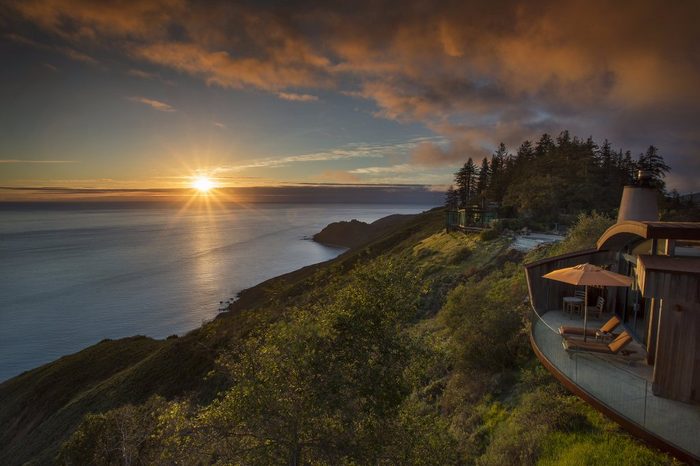 Sun sets over ocean, casting golden light on a hillside with trees and a house featuring a patio with an umbrella and lounge chair.