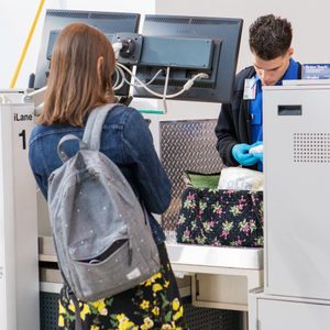 Security officer examines bag while traveler waits. They're at an airport security checkpoint, surrounded by screens and equipment. Sign reads, 