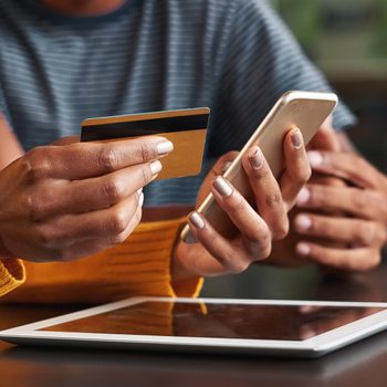Hands hold a credit card and smartphone, performing an online transaction on a tablet. A person in a striped shirt is nearby.