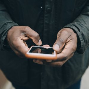 Hands holding a smartphone, typing a message, dressed in a dark jacket, against a neutral background.
