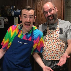 Two smiling people wear colorful aprons in a kitchen, with a refrigerator and wooden cabinets in the background. The aprons read 