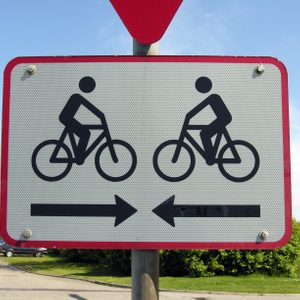 Sign depicts cyclists moving in opposite directions, accompanied by arrows pointing left and right, with a backdrop of greenery and sky.
