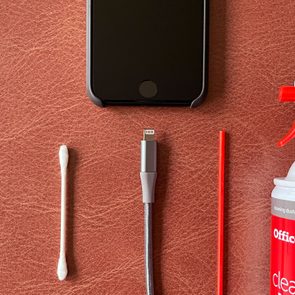 Phone and cleaning tools arranged neatly on brown leather surface, featuring cotton swab, cable, straw, and Office Depot cleaning duster.