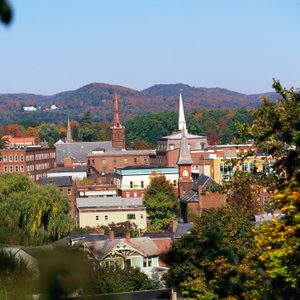 Buildings with steeples stand amidst trees in vibrant autumn colors, surrounded by hills under a clear blue sky.