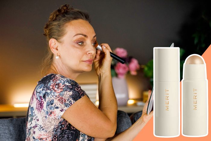 A woman applies makeup with a brush in a softly lit room; two Merit products are displayed on a peach background.