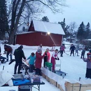 Children skate and play hockey on an outdoor rink, surrounded by adults and trees, near a red barn in a snowy setting.