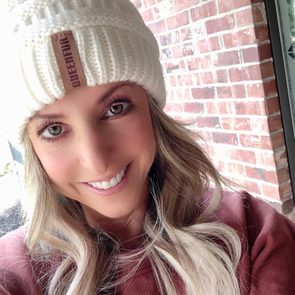 A person smiles, wearing a white knit hat labeled "QUEENFUR," in front of a red brick wall.