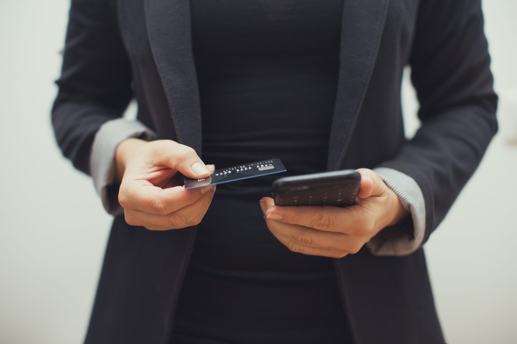 Person holds a credit card and smartphone, appearing to input information, while wearing a dark suit in a neutral, indoor setting.