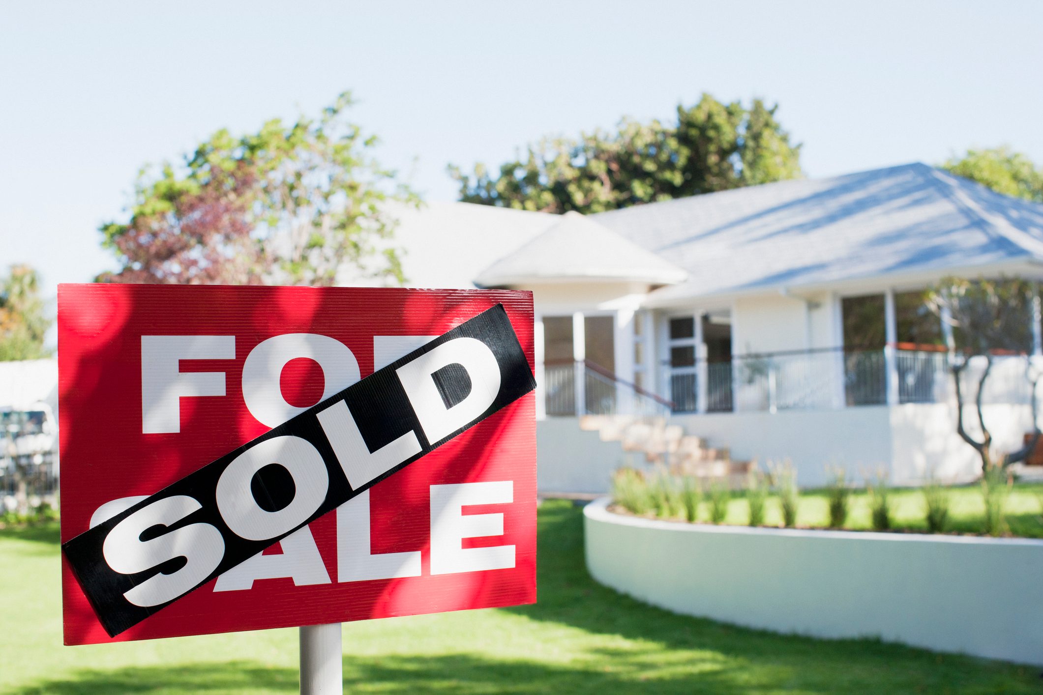 Sign reads "FOR SALE" with "SOLD" overlay; stands in front of a white house surrounded by a green lawn and trees.
