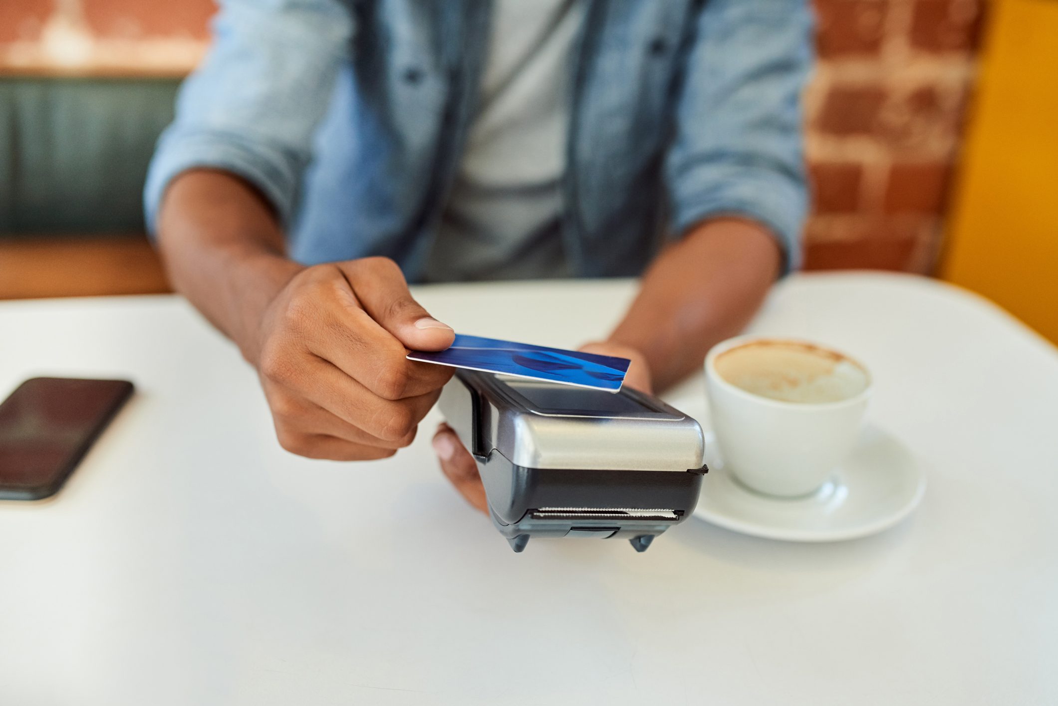 Hand swipes card on payment terminal, seated at a table. Nearby are a smartphone and a cup of coffee on a saucer.