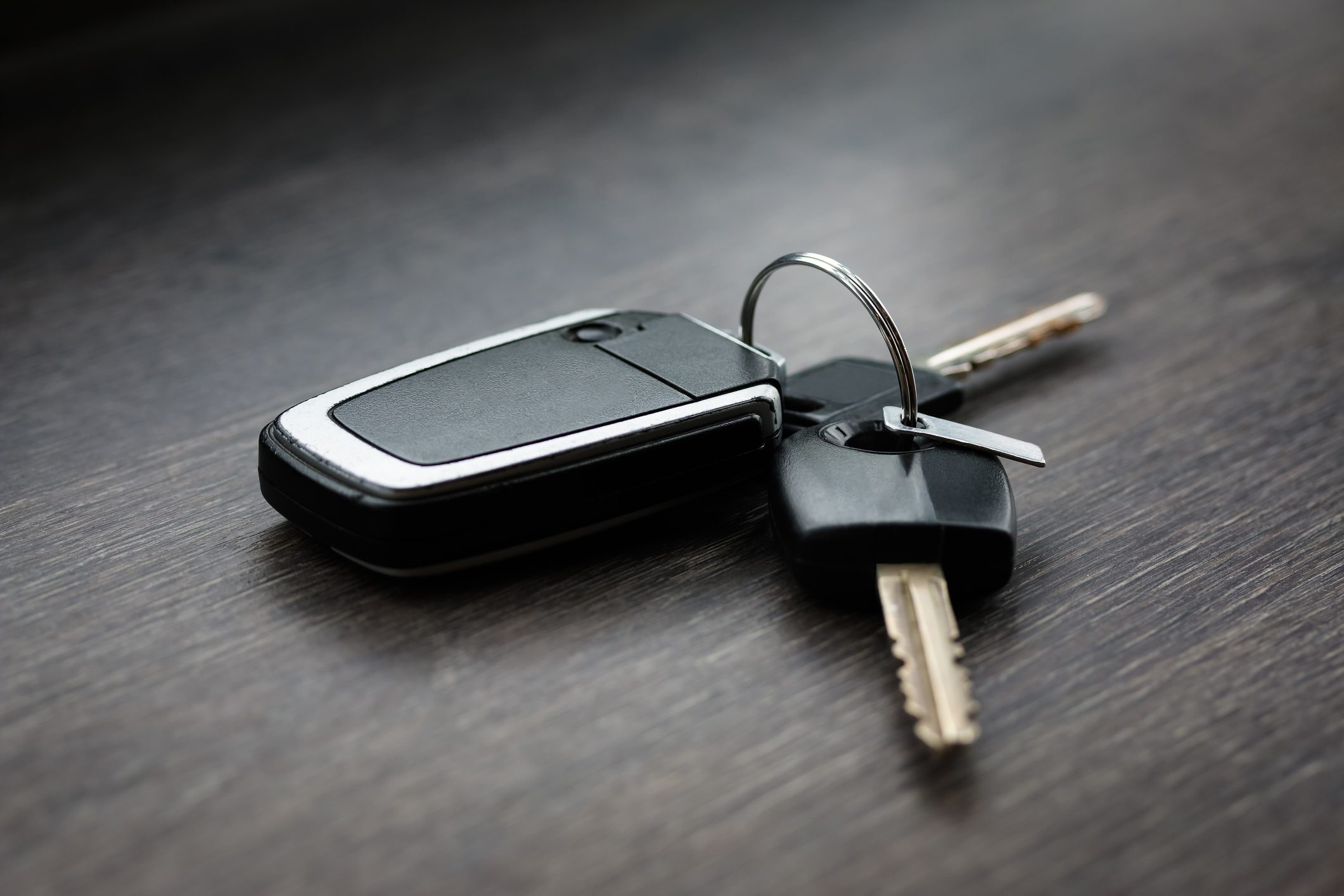 Car keys rest on a dark wooden surface, featuring a black plastic fob with metallic edges, attached to a key ring.