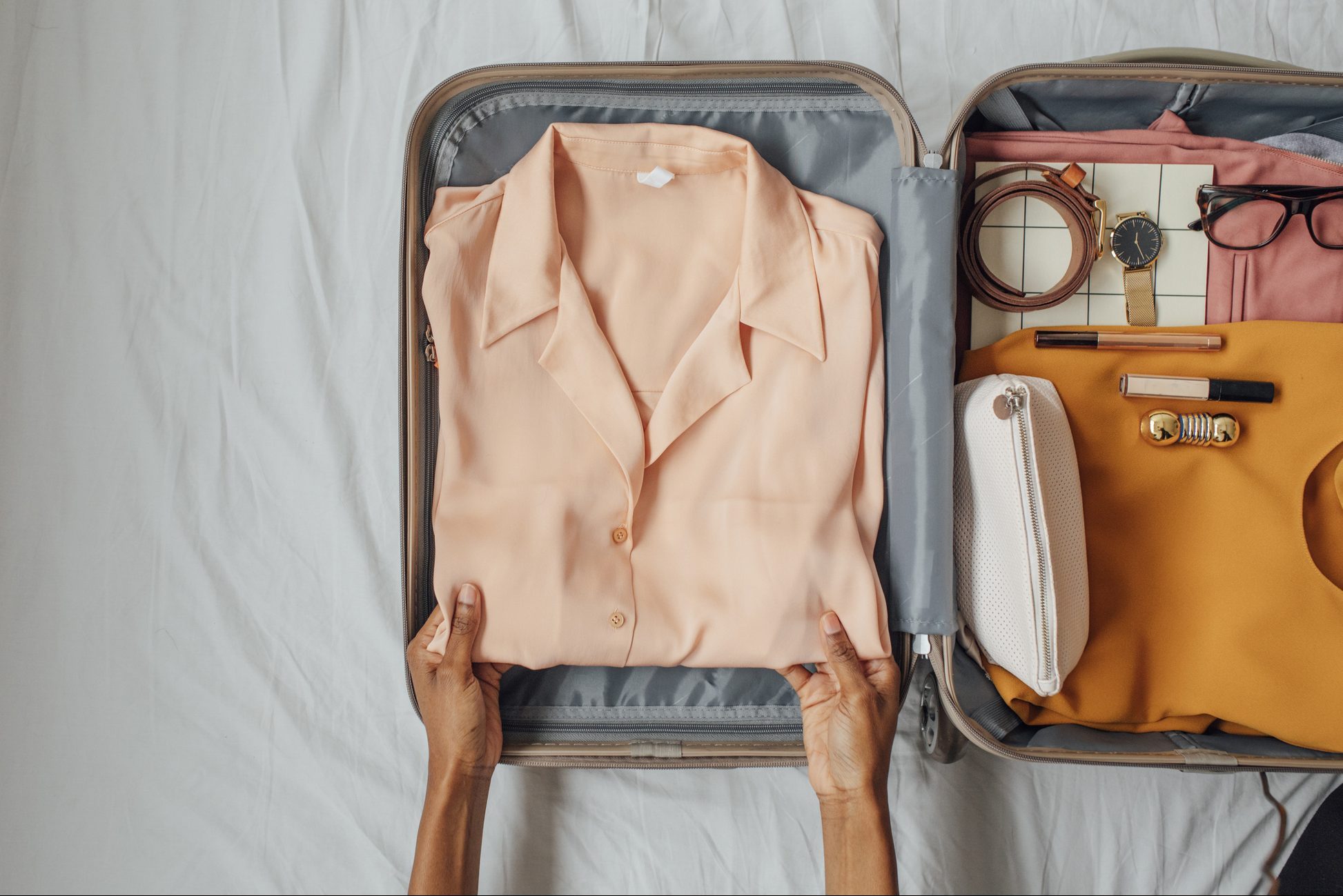 Hands fold a peach shirt inside a suitcase on a bed, surrounded by accessories like glasses, a watch, a belt, and cosmetic items.