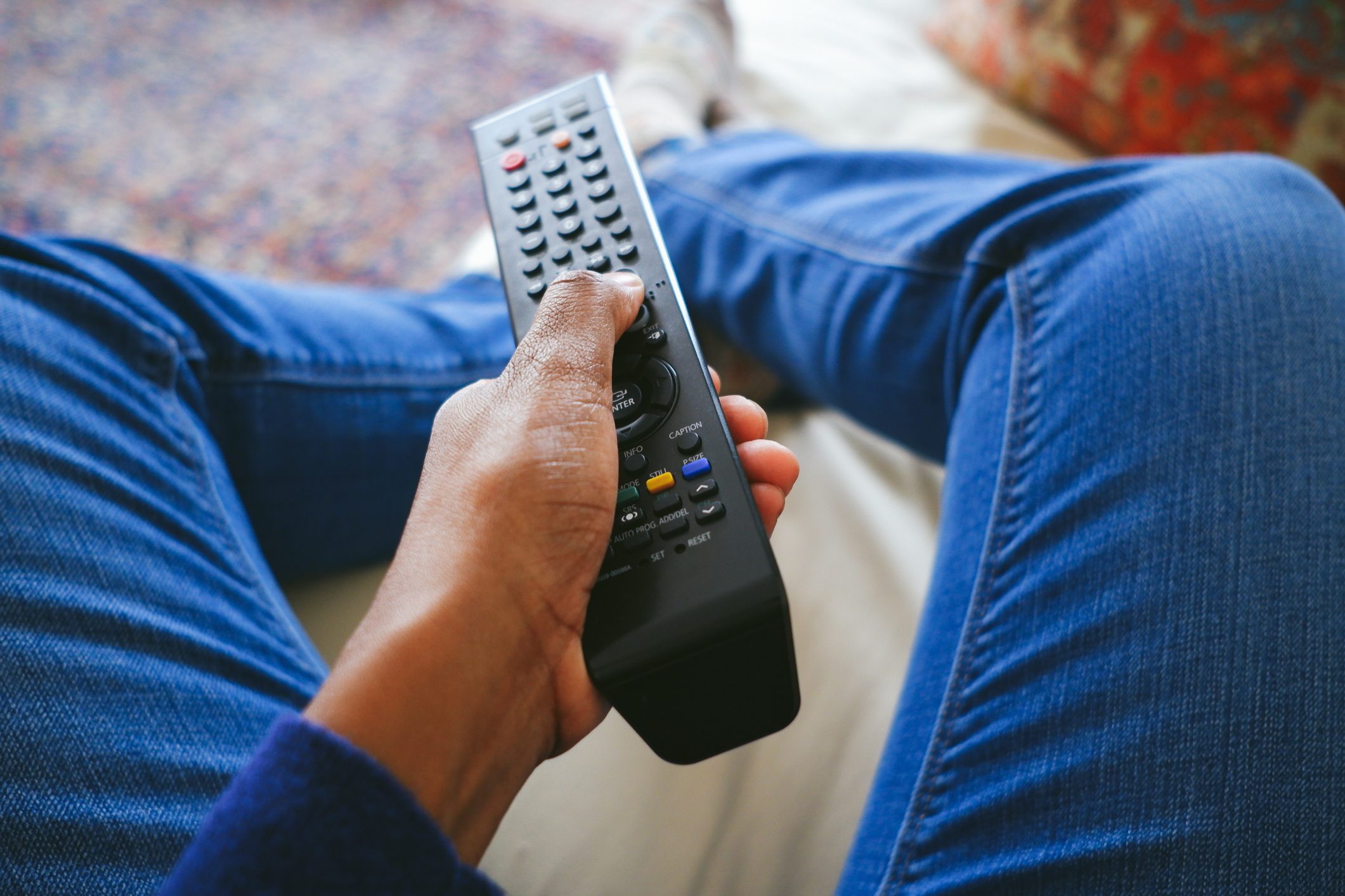 Hand holds a remote control, positioned over crossed legs in jeans, seated on a couch.