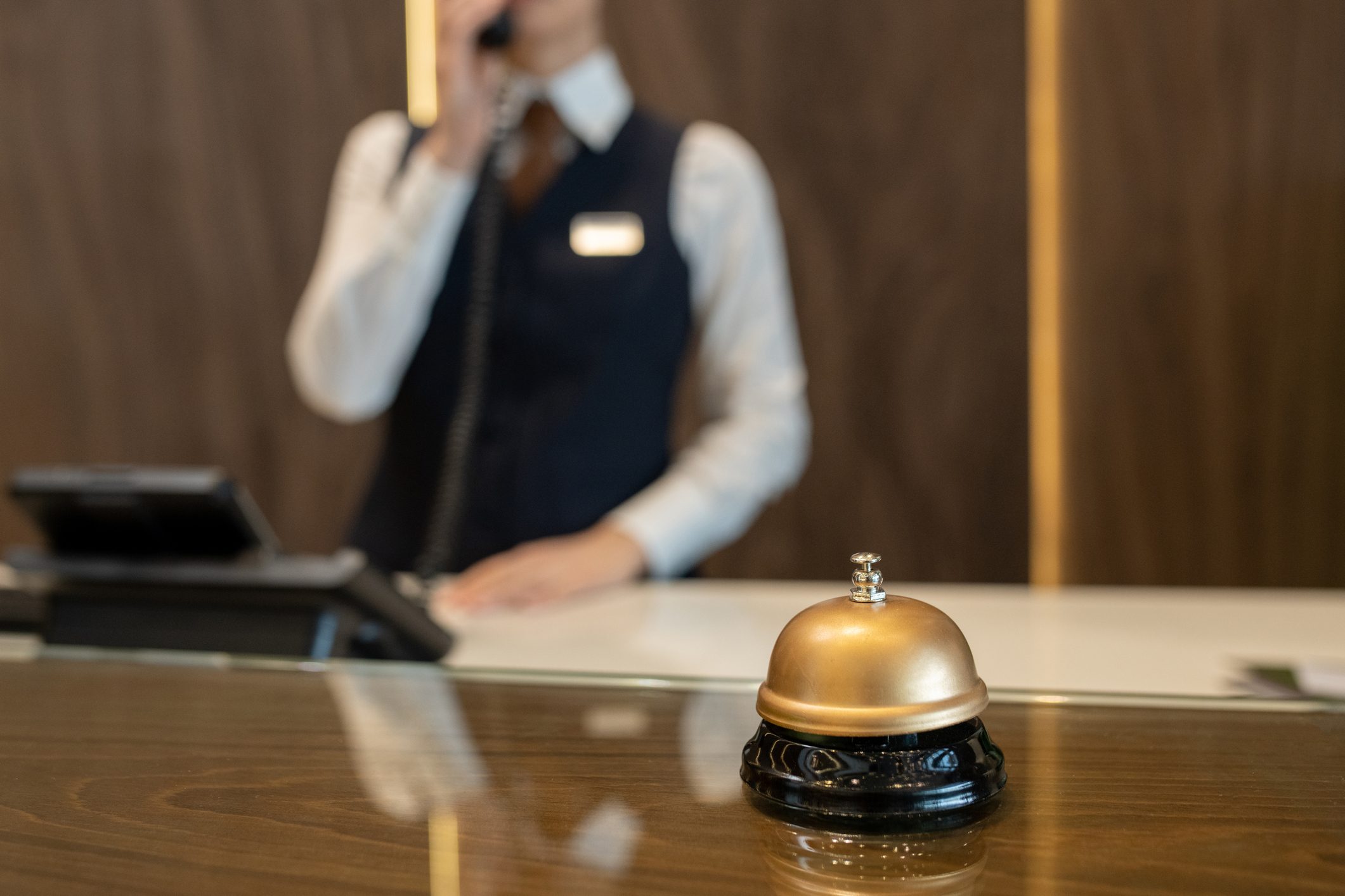 A hotel bell sits on a counter while a staff member speaks on the phone in the background.