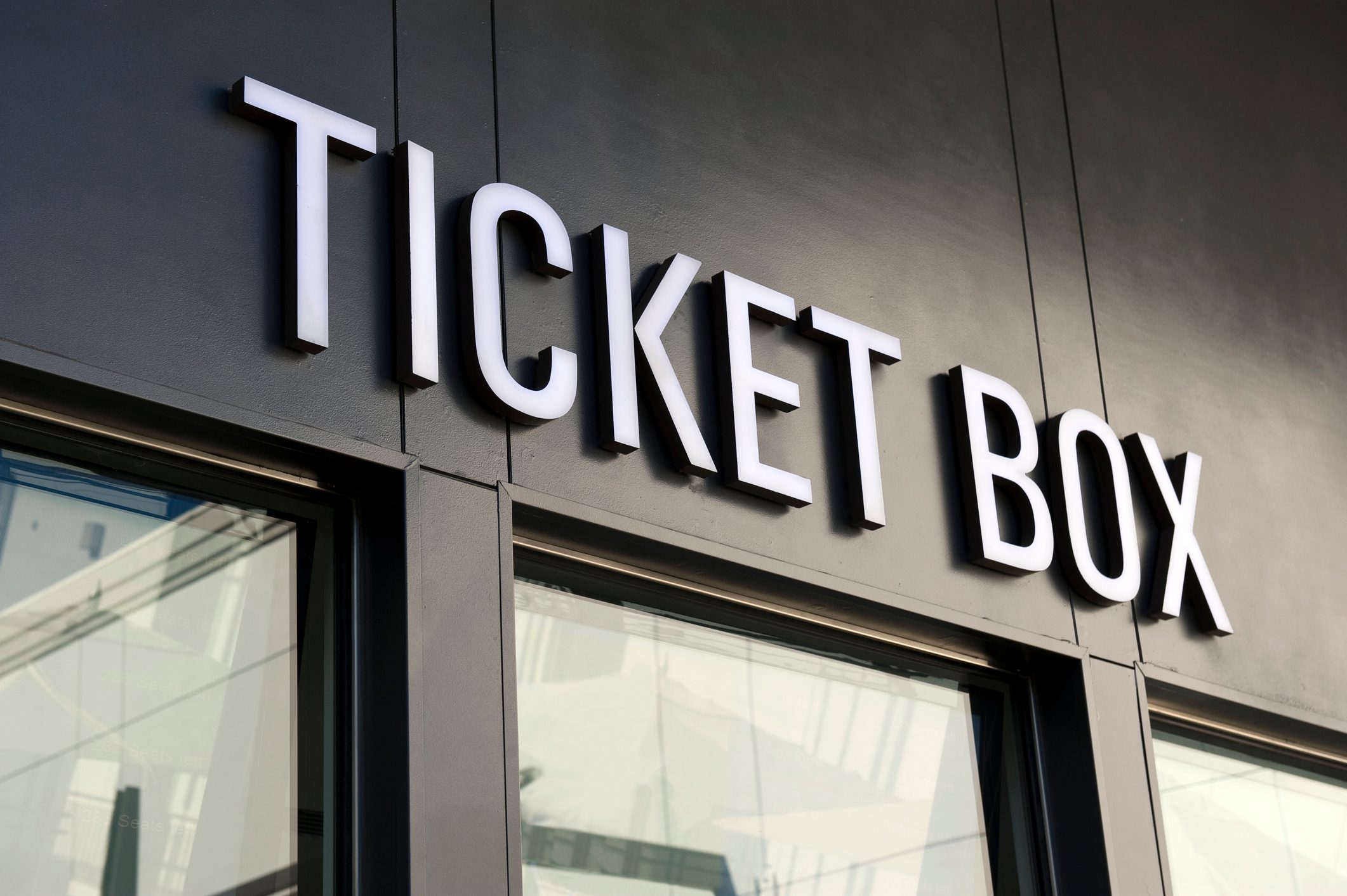 Sign displays "TICKET BOX" on a building's dark facade, positioned above windows reflecting a modern architectural setting.