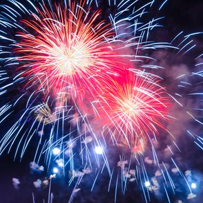 Fireworks burst vibrantly against a dark night sky, displaying red and blue streaks with smoke trails enhancing the festive atmosphere.
