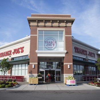 Storefront displays "TRADER JOE'S" logo; customers enter and exit amidst parked cars and shopping carts; sunny suburban setting.