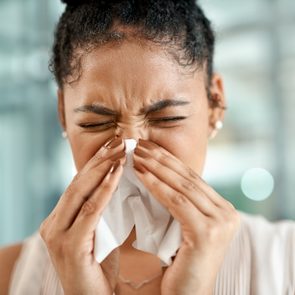 Person sneezes into tissue, eyes closed tightly, in a blurred indoor setting.