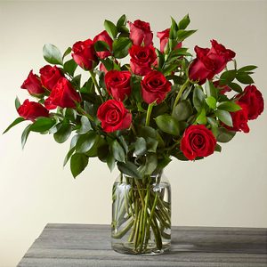 Red roses stand in a clear glass vase on a wooden table, surrounded by green leaves.