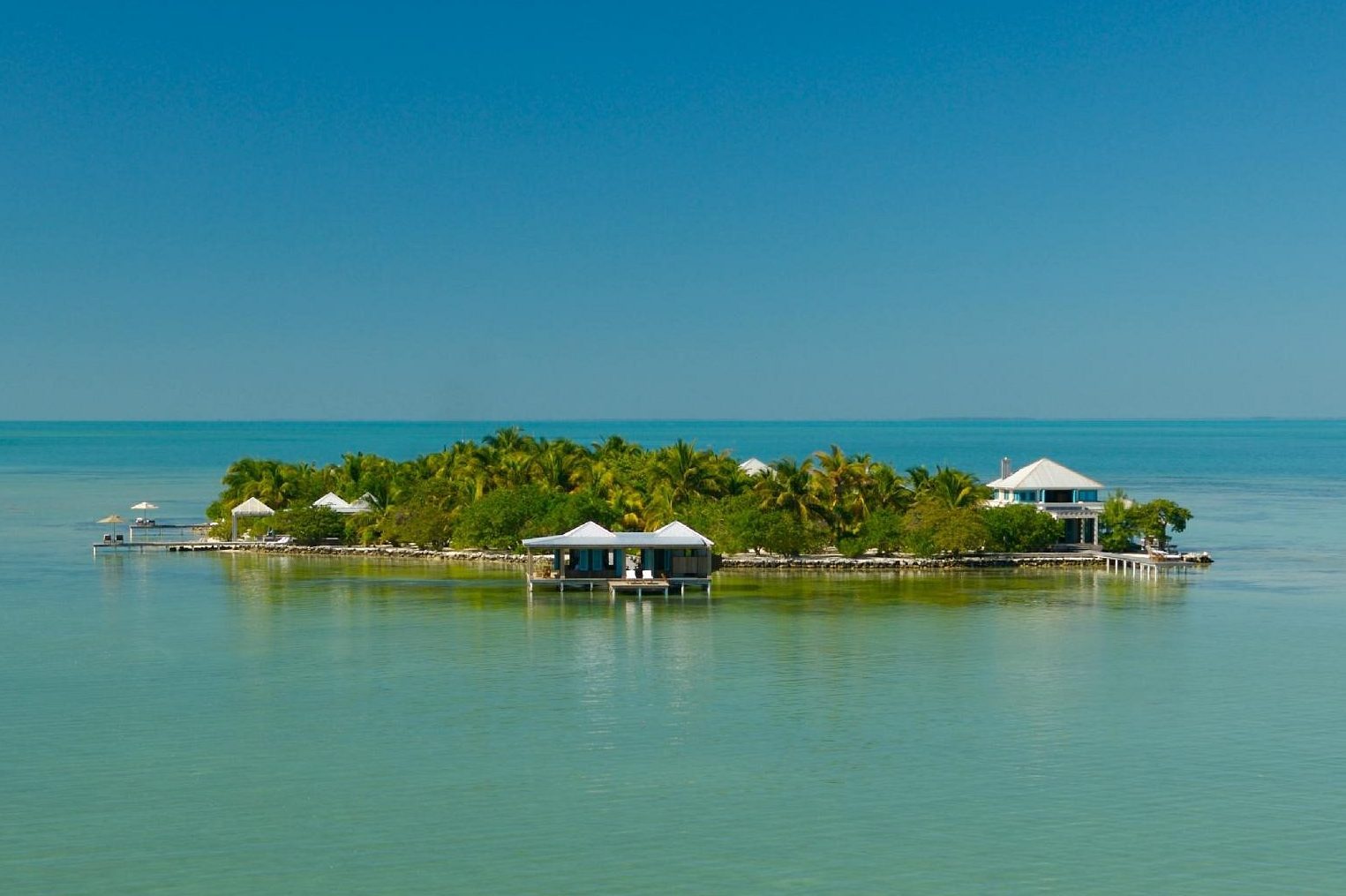 Island houses surrounded by lush palm trees sit peacefully amidst clear turquoise water under a bright blue sky.