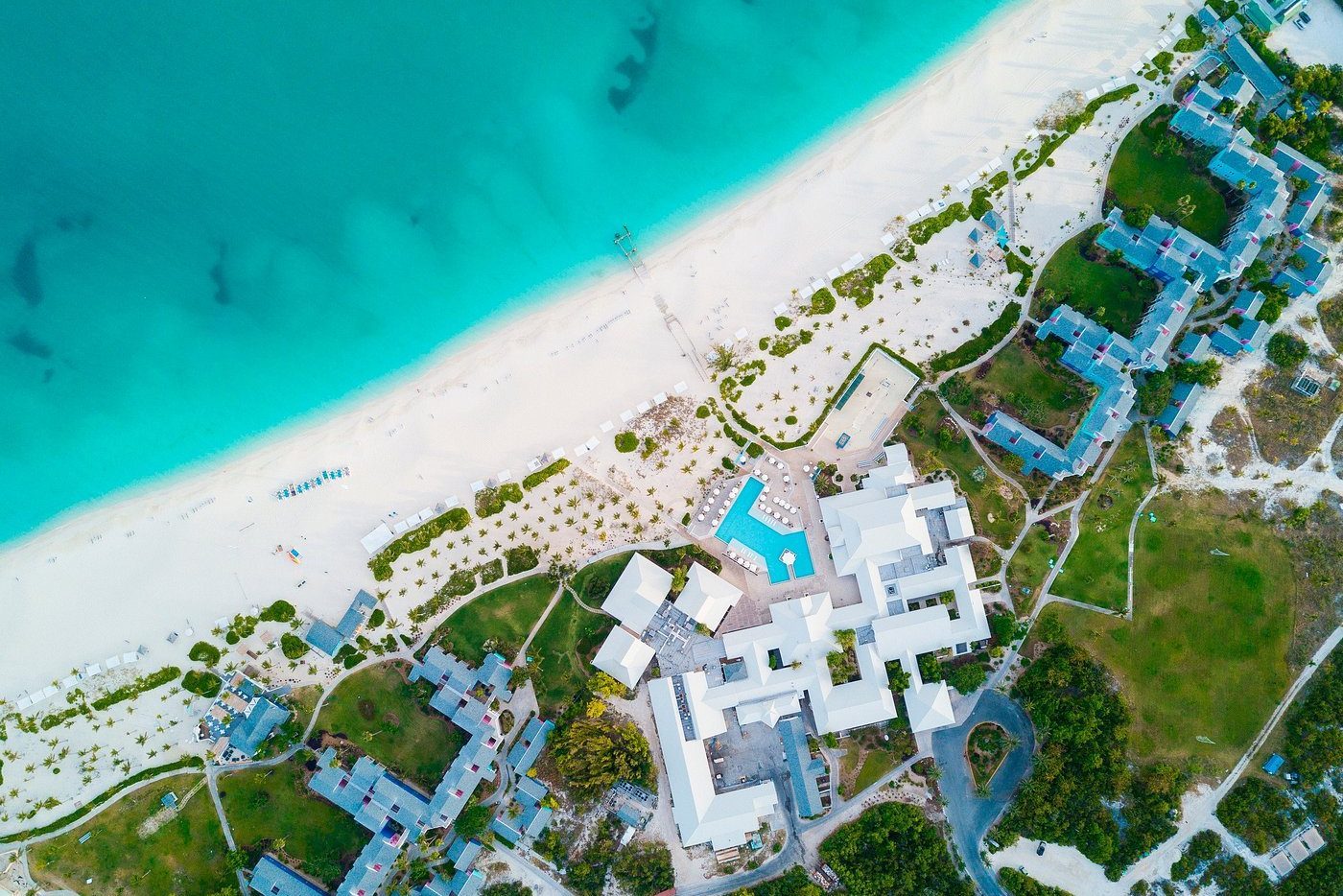 Beach resort buildings surround a central pool, bordered by sandy shores and turquoise sea, viewed from above.