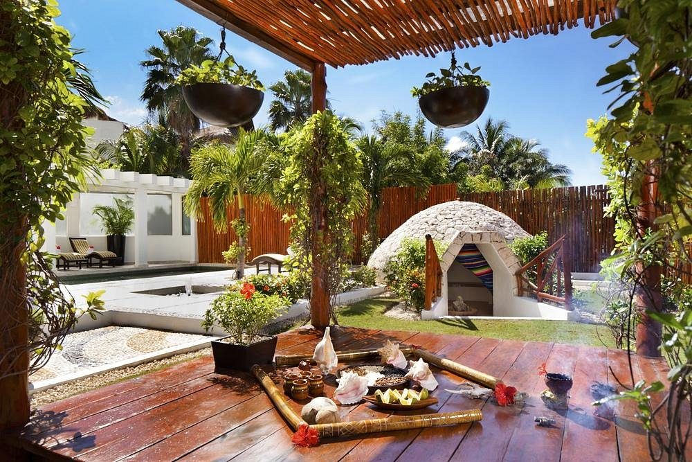 A wooden deck displays pottery and plants under a pergola, with a stone hut amidst lush greenery in a sunlit tropical garden.