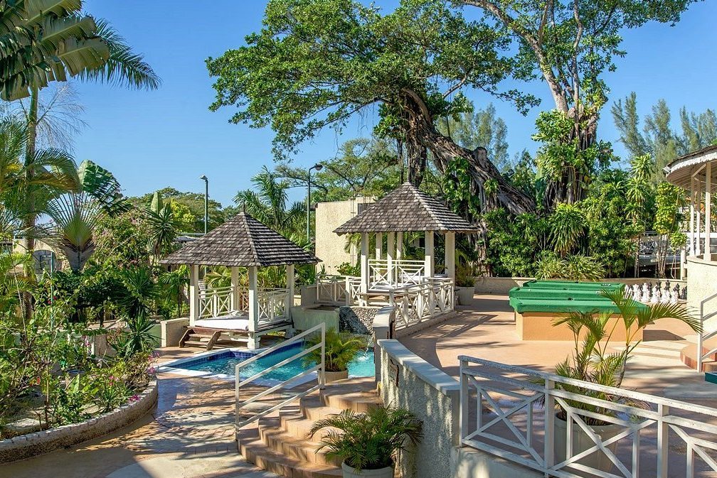 Gazebo stands over a small pool, surrounded by lush tropical plants and trees, under a clear blue sky.