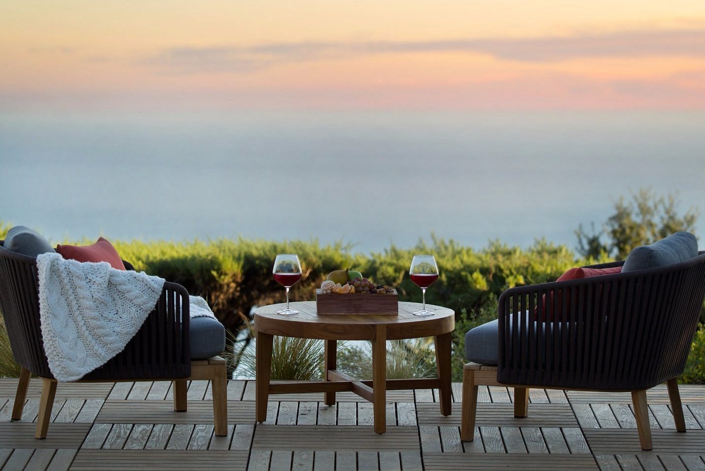 Two armchairs face a wooden table with wine glasses and snacks, set against a serene ocean sunset.
