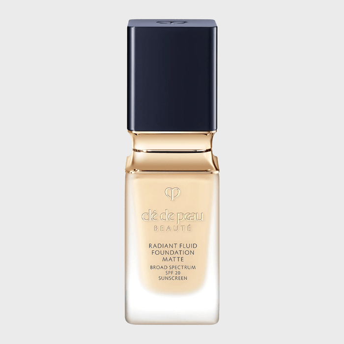 Bottle displays makeup foundation, standing upright, with a black cap and gold accents on a plain gray background. Text reads 