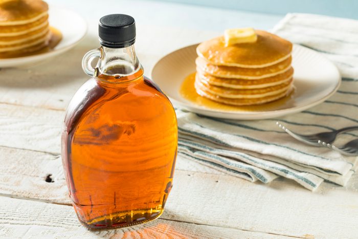 Bottle of amber syrup sits on table; pancakes with butter on a plate in the background, atop striped cloth.
