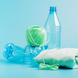 Green yarn rests on crushed blue plastic bottles, accompanied by a folded mint-green fabric, all against a light blue background.