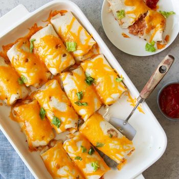 Burritos topped with melted cheese are arranged in a baking dish; one is on a plate. Salsa sits beside on a gray countertop.
