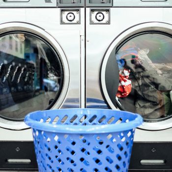 Front-load washing machines, with visible clothes inside, sit in a laundromat setting, accompanied by a blue laundry basket in the foreground.