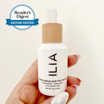 Hand holds a white serum bottle with a tan cap; "ILIA Super Serum Skin Tint SPF 40" is printed. Reader's Digest badge shown nearby.