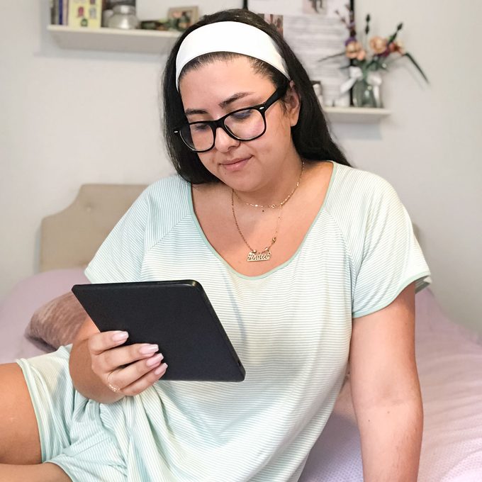 A person reads an e-reader while sitting on a bed in a softly decorated bedroom with shelves and flowers.
