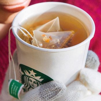 Hands hold a warm Starbucks cup with tea bags inside, against a cozy red sweater background.